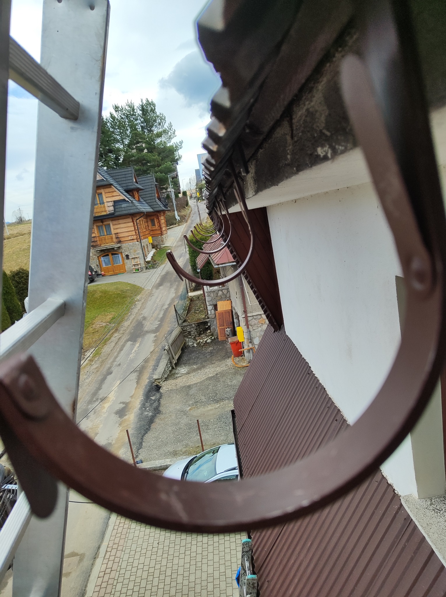 Widok z drabiny na rynny dachowe, fragment dachu z blachy trapezowej, ulicę z domami w stylu góralskim i samochód. Ujęcie z góry, z perspektywy osoby pracującej przy dachu.