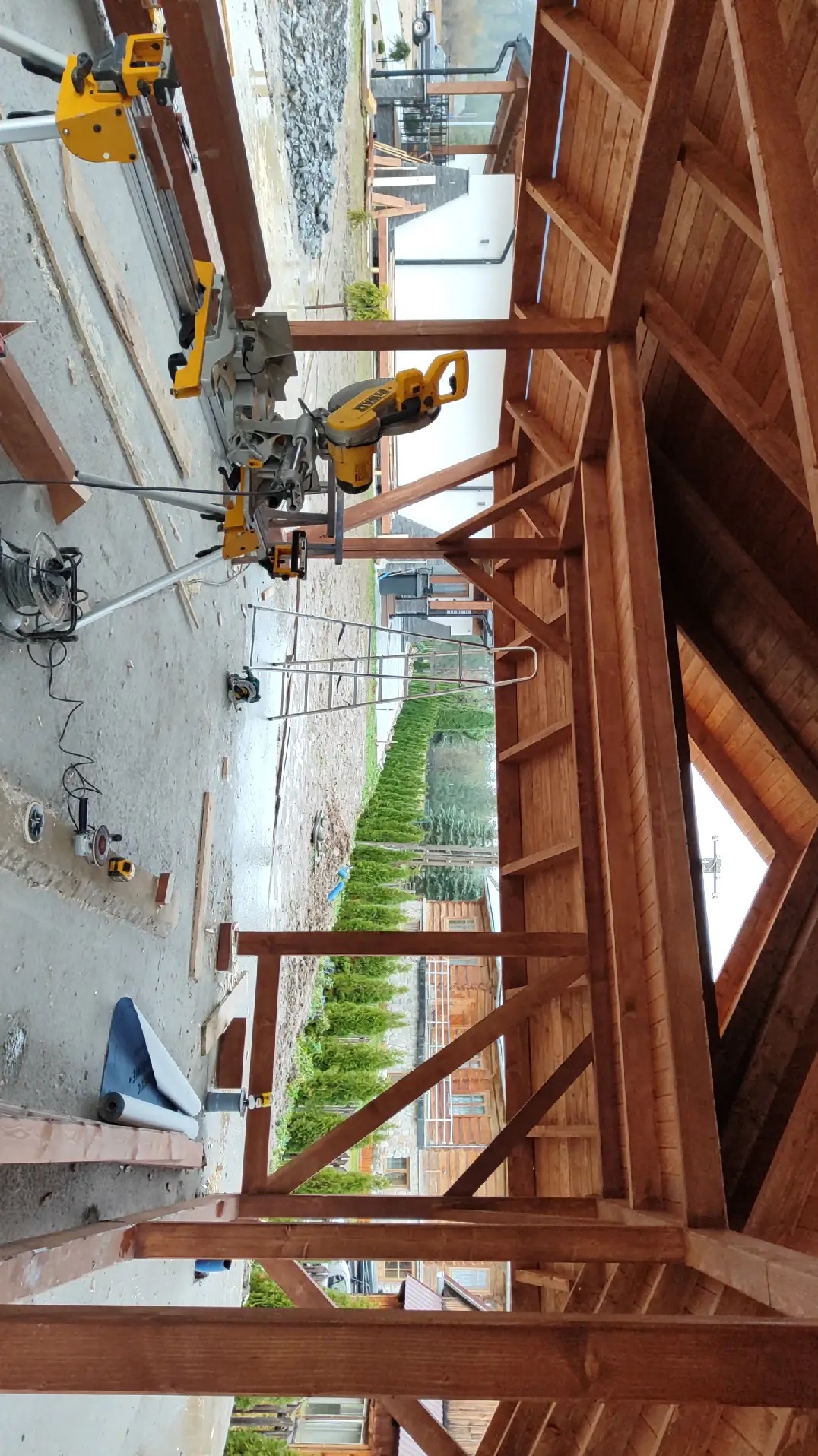 Drewniana konstrukcja dachu w trakcie budowy, widok od wewnątrz, na zewnątrz widoczny ogród i domy, w tle narzędzia budowlane i drabina.