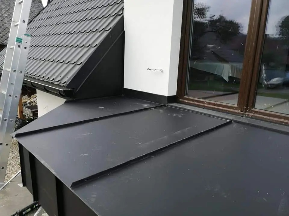 Szczegółowe ujęcie nowej, czarnej, metalowej obróbki blacharskiej przy oknie dachowym, z widoczną drabina opartą o dach z dachówki ceramicznej.