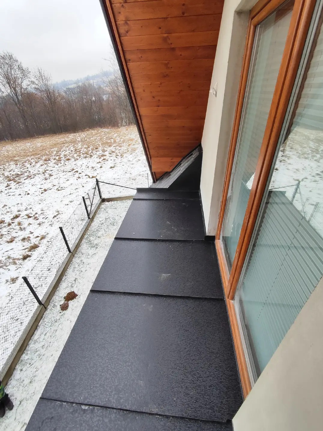 Nowy, czarny dach z blachy na budynku jednorodzinnym, widok z góry w zimowy dzień, widoczne drewniane podbitki i okno z drewnianą ramą, na zewnątrz ośnieżone pole i siatka ogrodzeniowa.