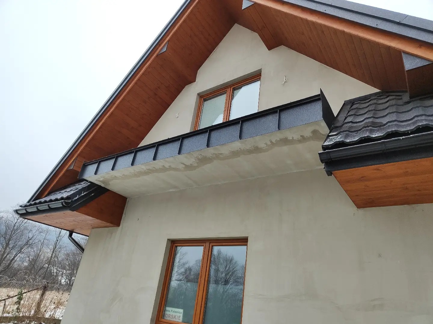 Fragment elewacji domu jednorodzinnego z widocznymi drewnianymi elementami więźby dachowej, czarnymi obróbkami blacharskimi i rynnami. Ściana w trakcie wykańczania, widoczne ślady po pracach...