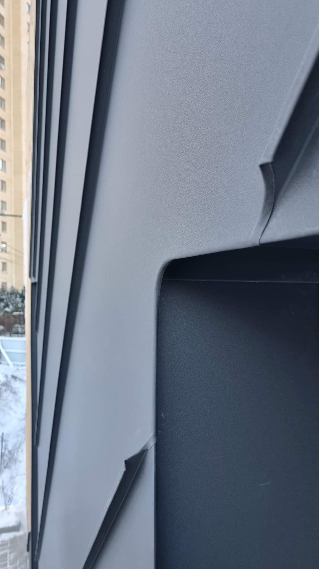 Szczegółowe ujęcie szarej blachy na elewacji budynku z widocznymi pionowymi łączeniami paneli i fragmentem okna w tle.