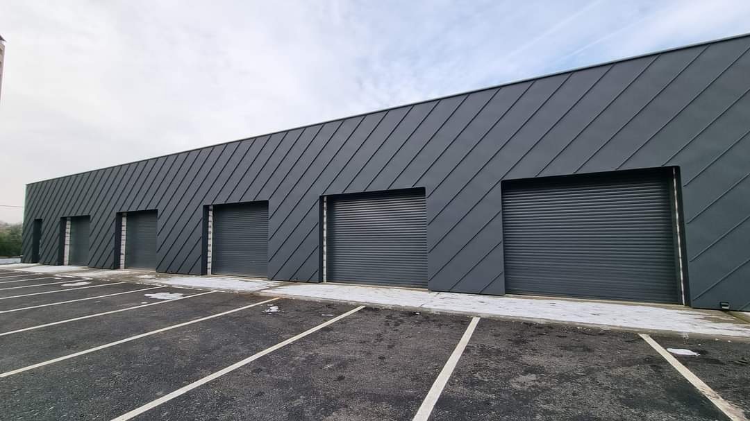 Szara hala przemysłowa z ukośną blachą na elewacji i czterema bramami rolowanymi, widok z parkingu z wyznaczonymi miejscami.