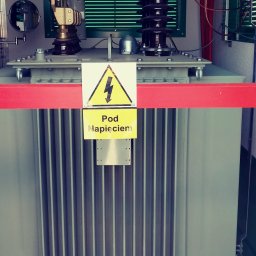 ProElektroeM Michał Zychowicz - Transformator energetyczny z żółtą tabliczką ostrzegawczą 'Pod Napięciem', zabezpieczony czerwoną barierką, widoczne kable i elementy instalacji elektrycznej.