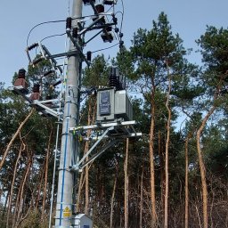 ProElektroeM Michał Zychowicz - Betonowy słup energetyczny z zamontowanym transformatorem, skrzynką elektryczną i osprzętem, otoczony lasem sosnowym, widoczne oznaczenia ostrzegawcze.