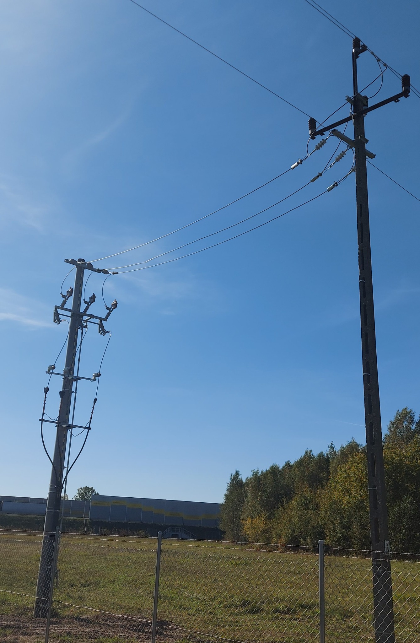 Dwa słupy energetyczne połączone przewodami na tle błękitnego nieba, widok zza metalowego ogrodzenia, w tle drzewa i budynek.