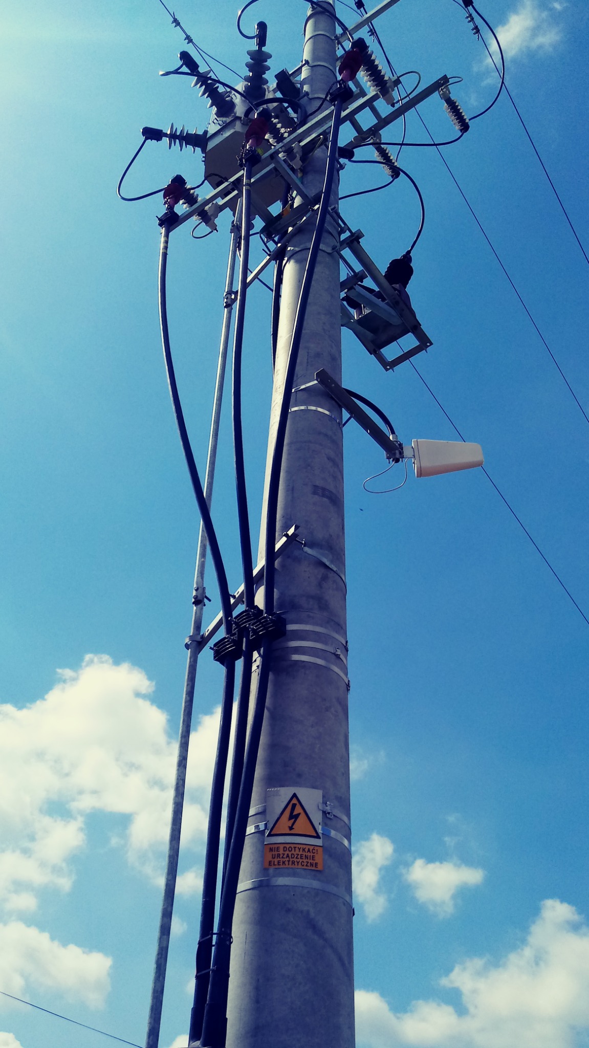 Betonowy słup energetyczny z transformatorem i przewodami na tle błękitnego nieba z obłokami w Sieradzu. Widoczna tabliczka ostrzegawcza o urządzeniach elektrycznych.