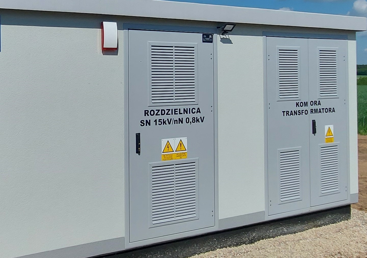 Zewnętrzna stacja transformatorowa z dwiema szarymi szafami: Rozdzielnica SN 15kV/nN 0,8kV i Komora Transformatora, zabezpieczona lampą nad drzwiami, z widocznym znakiem ostrzegawczym.