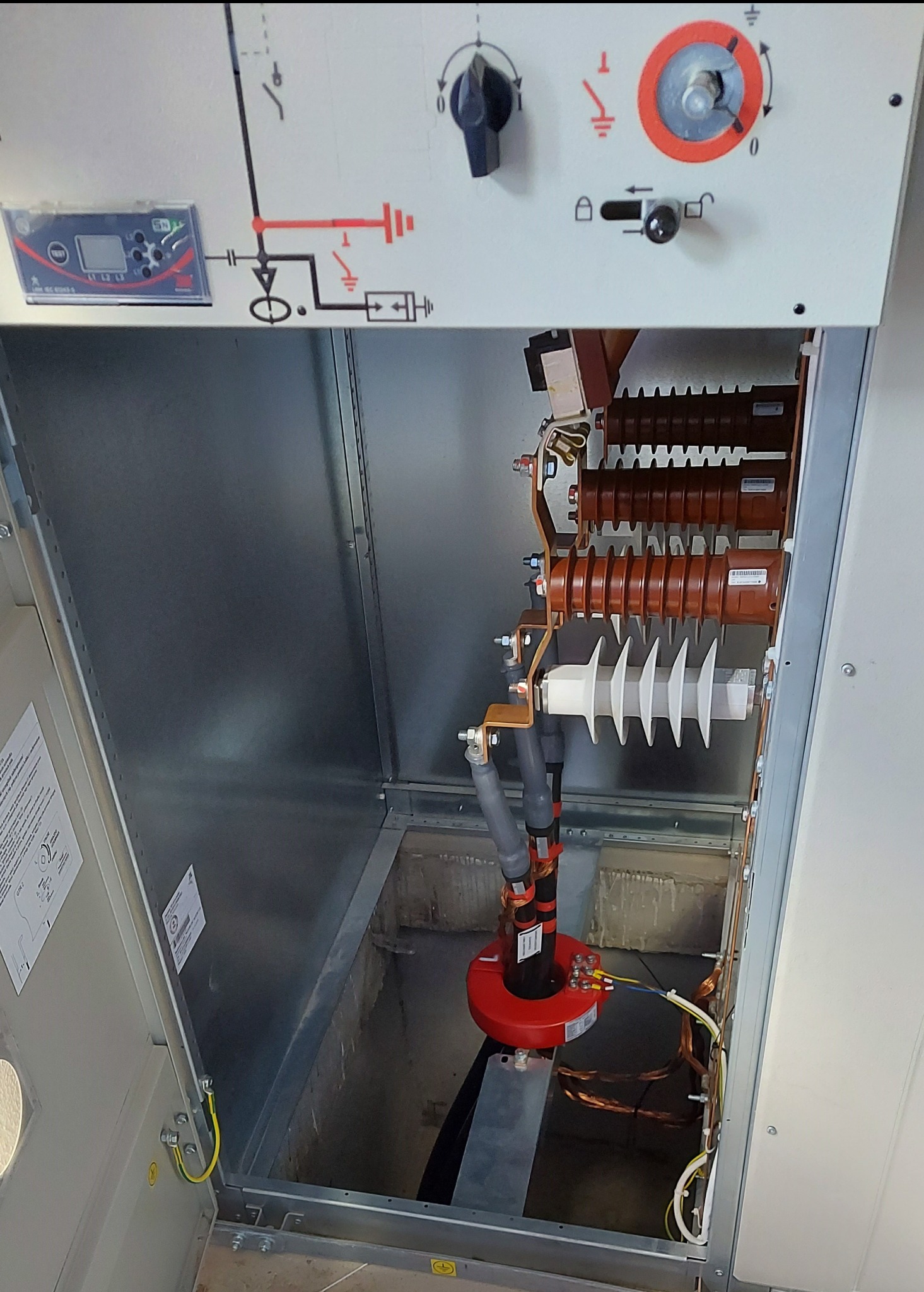 Wnętrze rozdzielnicy elektrycznej średniego napięcia z widocznymi izolatorami, przewodami miedzianymi i elementami sterującymi na panelu przednim.