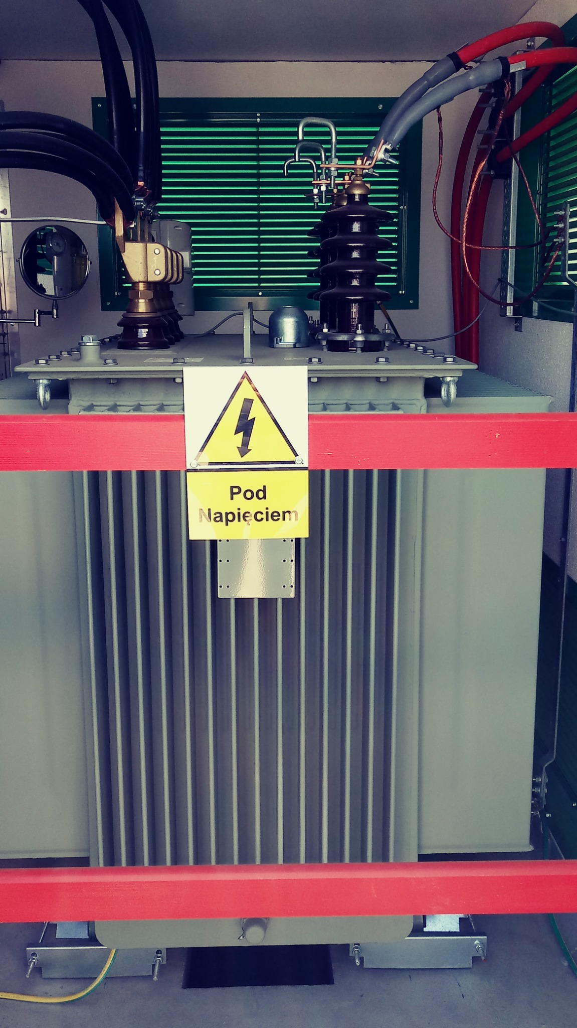 Transformator energetyczny z żółtą tabliczką ostrzegawczą 'Pod Napięciem', zabezpieczony czerwoną barierką, widoczne kable i elementy instalacji elektrycznej.