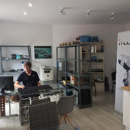 Itludek Michał Osuch - Mężczyzna w okularach siedzi przy szklanym biurku w biurze IT, na półkach widoczne komponenty komputerowe, w tle baner reklamowy z logo firmy i wizerunkiem robota.