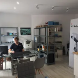 Mężczyzna w okularach siedzi przy szklanym biurku w biurze IT, na półkach widoczne komponenty komputerowe, w tle baner reklamowy z logo firmy i wizerunkiem robota.