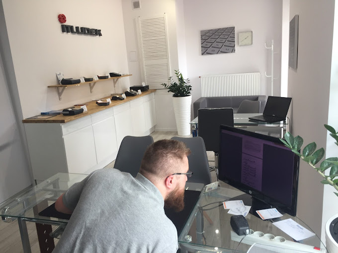 Mężczyzna z brodą siedzi przy szklanym biurku, pracując na komputerze w jasnym biurze z logo 'MUDEK' na ścianie i półkami z urządzeniami; laptop na drugim biurku w tle.