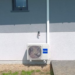 MCSerwisPlus - Biała jednostka zewnętrzna klimatyzacji Haier zamontowana na elewacji domu z widoczną instalacją odprowadzającą skropliny i fragmentem węża ogrodowego na trawie.