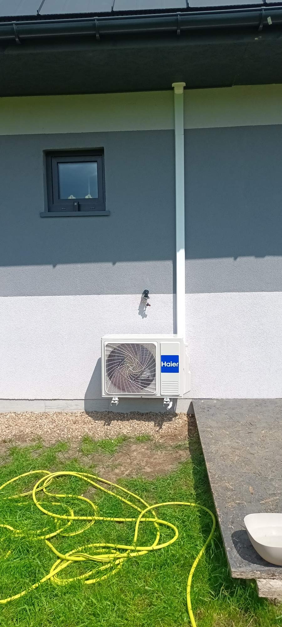 Biała jednostka zewnętrzna klimatyzacji Haier zamontowana na elewacji domu z widoczną instalacją odprowadzającą skropliny i fragmentem węża ogrodowego na trawie.