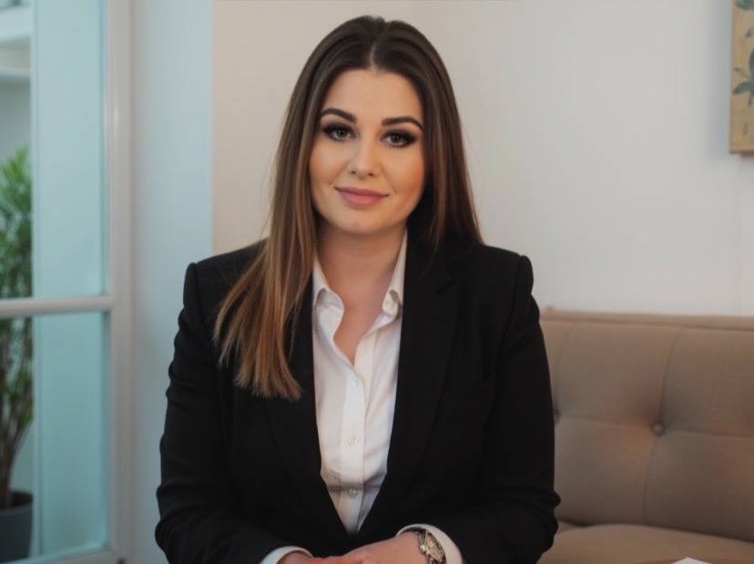 Portret kobiety w czarnym żakiecie i białej koszuli, siedzącej na beżowej sofie. Uśmiechnięta, z delikatnym makijażem i długimi włosami. W tle okno i obraz.