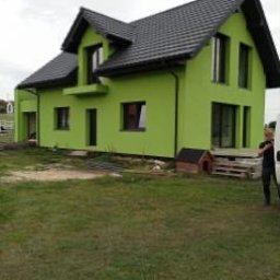 REBLOOM KRZYSZTOF KWIATUSZEWSKI-ZAJĄC - Nowoczesny dom jednorodzinny z zieloną elewacją i czarnym dachem, widok z boku na trawnik, obok budynek gospodarczy i palety.