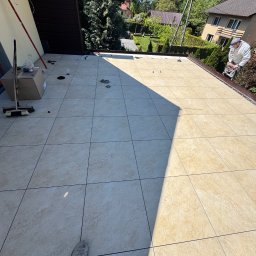 REBLOOM KRZYSZTOF KWIATUSZEWSKI-ZAJĄC - Jasne płytki na tarasie, widoczne narzędzia i pracownik docinający element. Tło: domy i zieleń. Dzień słoneczny, cień na płytkach. Widok z góry, stopa w kadrze.