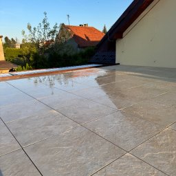 REBLOOM KRZYSZTOF KWIATUSZEWSKI-ZAJĄC - Wyłożony jasnoszarymi płytkami taras z widokiem na domy i zieleń w słoneczny dzień. Płytki odbijają światło, tworząc efekt połysku. Widoczny fragment elewacji budynku.