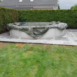 REBLOOM KRZYSZTOF KWIATUSZEWSKI-ZAJĄC - Okrągły basen ogrodowy z szarą plandeką, otoczony nową, jasną, betonową posadzką z regularnych płyt, obok zielony trawnik i wysokie tuje.