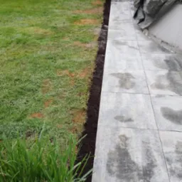 Ułożone płyty tarasowe z szarego betonu, widoczne świeże obrzeże z ziemi, trawa i zieleń w tle. Część tarasu przykryta plandeką. Widok z poziomu gruntu.