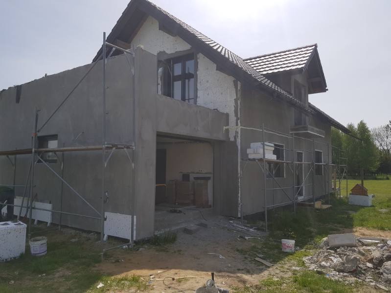 Dom w trakcie ocieplania elewacji: widoczne rusztowania, częściowo otynkowane ściany, styropian na fragmentach elewacji, nieporządek na posesji.