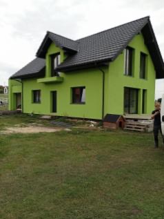 Nowoczesny dom jednorodzinny z zieloną elewacją i czarnym dachem, widok z boku na trawnik, obok budynek gospodarczy i palety.