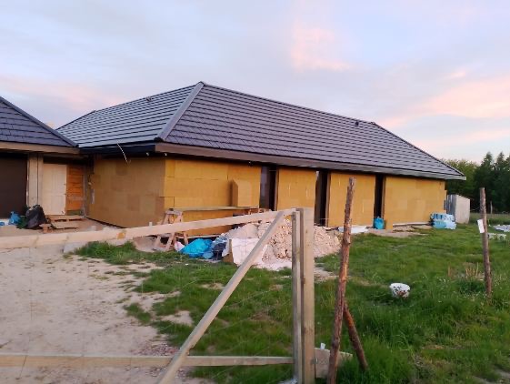 Dom jednorodzinny w trakcie prac: szara dachówka ceramiczna, ściany zewnętrzne pokryte żółtą izolacją, widoczne rusztowania i materiały budowlane na posesji, niebo o zmierzchu.