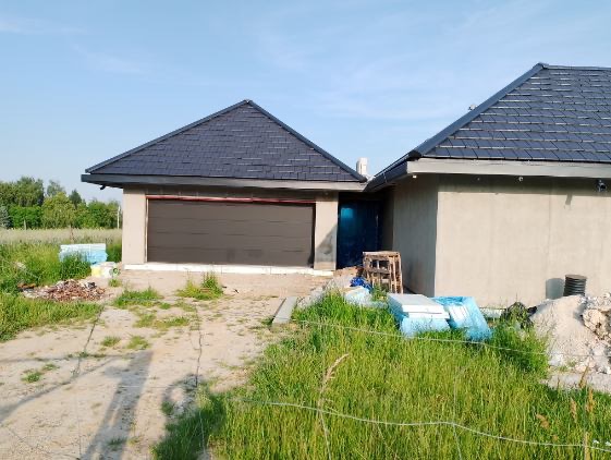 Nowy dom w trakcie budowy z ciemnoszarym dachem z dachówki bitumicznej i garażem z brązową bramą segmentową, widoczne materiały budowlane i nieotynkowana elewacja.
