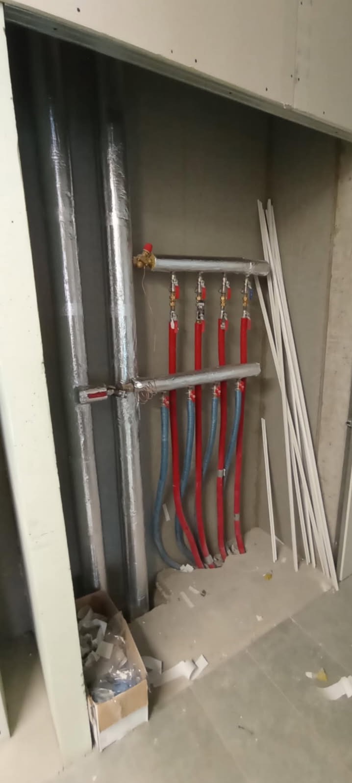 Rozdzielacz instalacji centralnego ogrzewania z rurami doprowadzającymi w kolorach czerwonym i niebieskim, zamontowany w surowym pomieszczeniu z widocznymi elementami konstrukcyjnymi i materiałami...