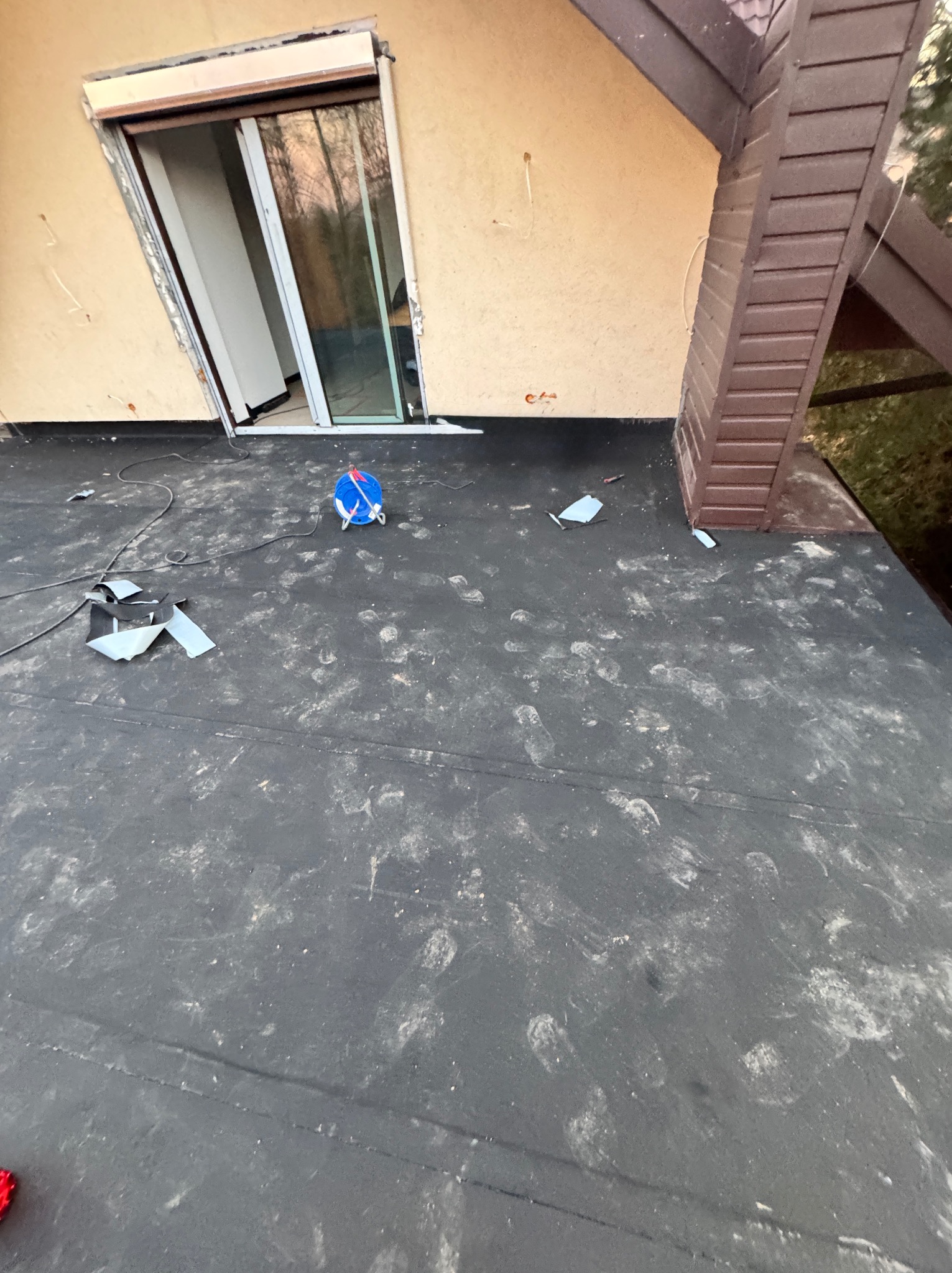 Czarny dach pokryty śladami butów, z otwartym oknem balkonowym i widocznym fragmentem elewacji oraz elementem konstrukcji dachu.