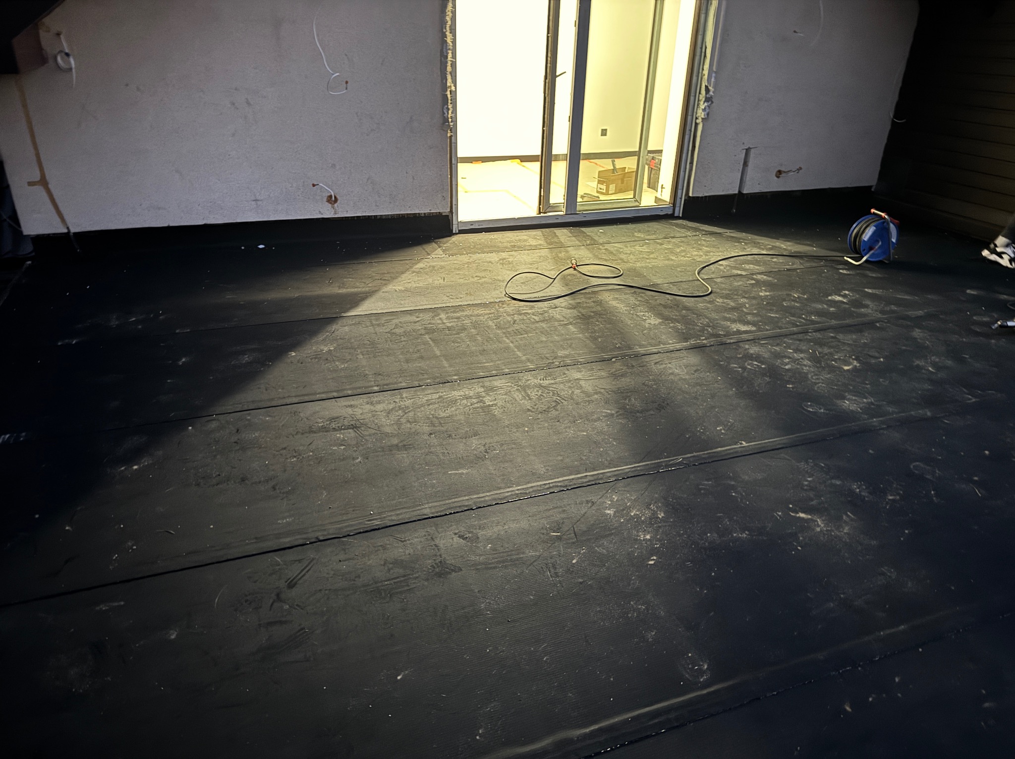 Czarna, gumowa hydroizolacja na balkonie, widoczna zwinięta rolka kabla, surowe ściany i otwarte drzwi balkonowe z widokiem na wnętrze.