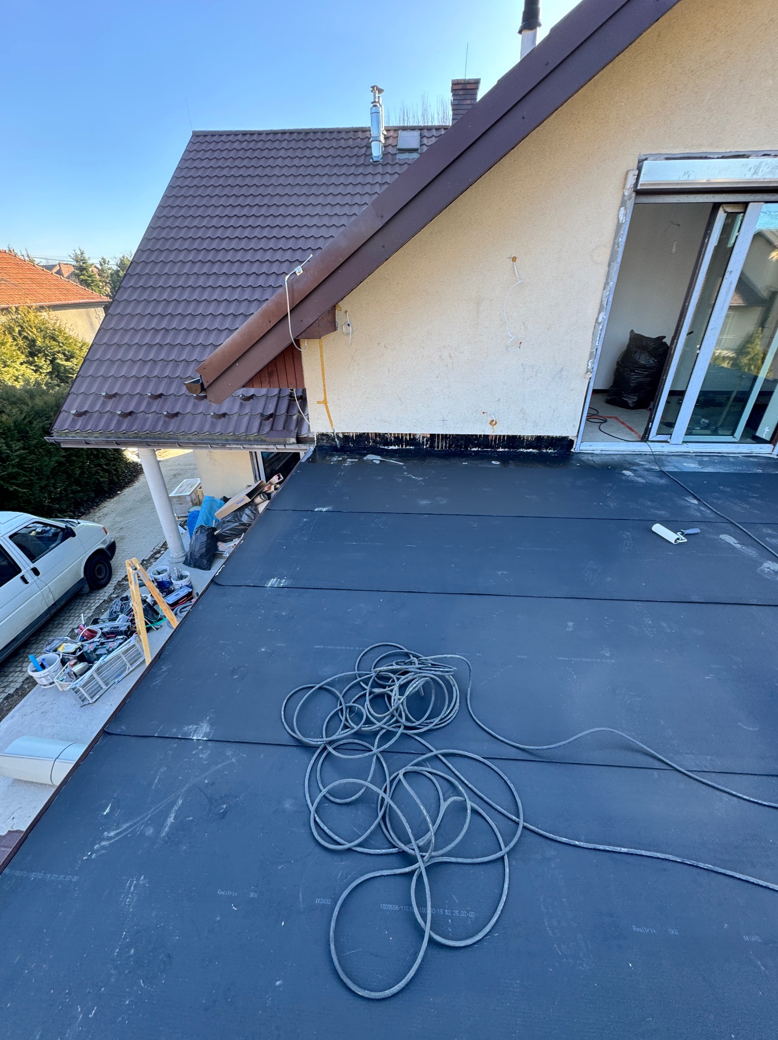 Czarna membrana dachowa z widocznymi zwojami kabli, w tle fragment dachu z brązową dachówką, ściana budynku z oknem balkonowym i pracownik w stroju roboczym.