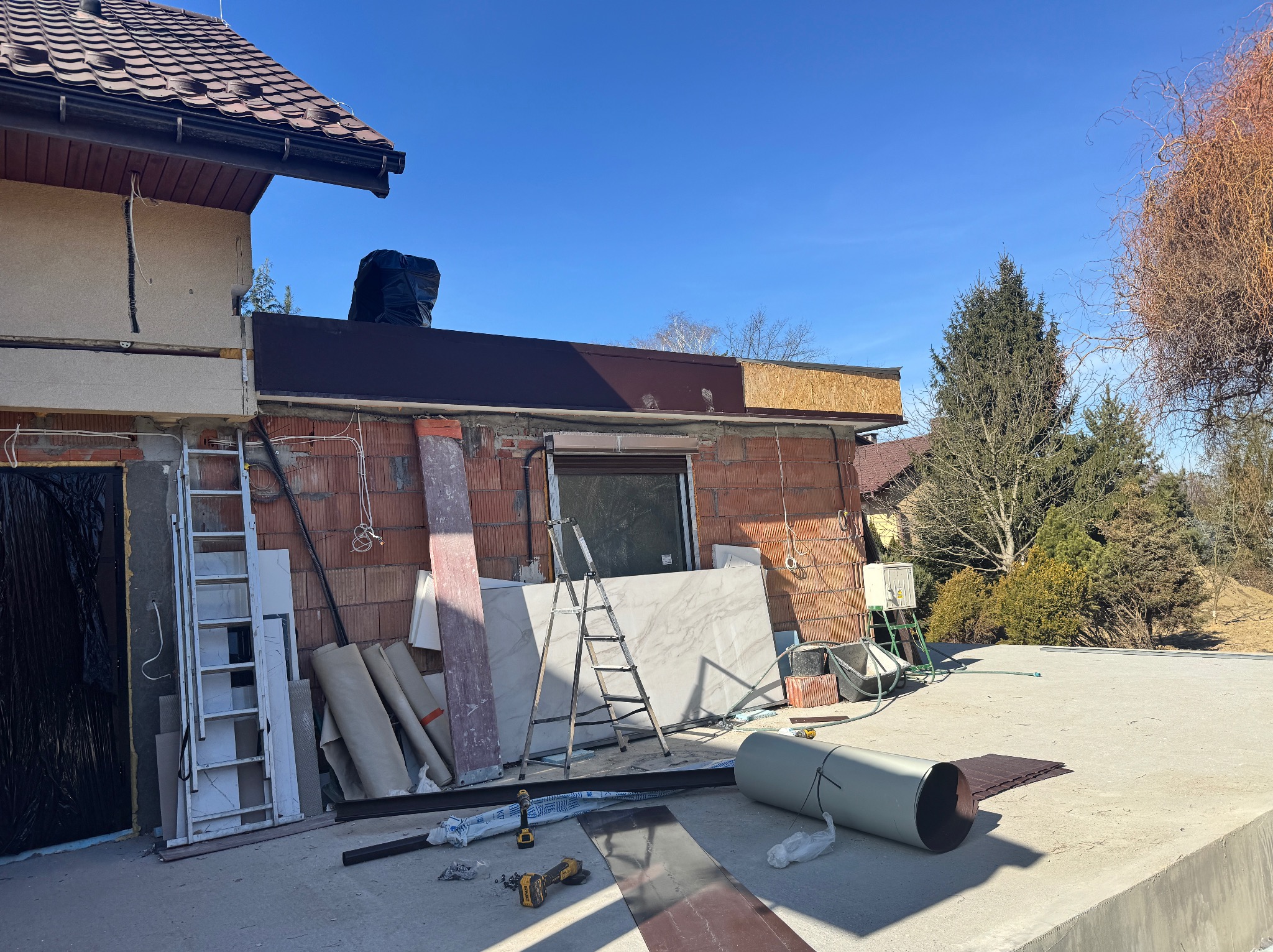 Budynek w trakcie budowy z widoczną czerwoną cegłą, elementami dachu i materiałami budowlanymi, w tym rolkami izolacji i płytami marmurowymi, z drabinkami i narzędziami na placu budowy w słoneczny...