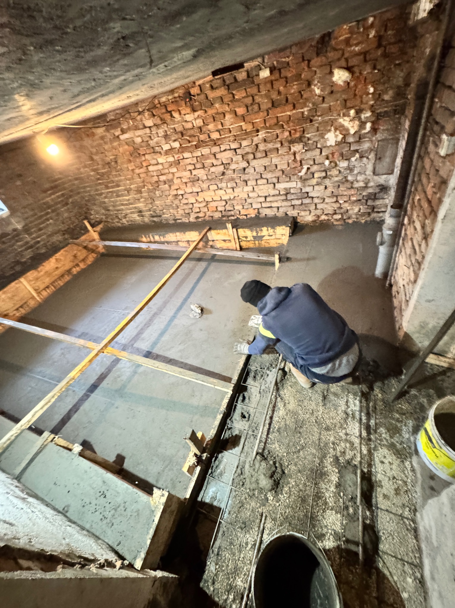 Robotnik w bluzie i czapce wyrównuje świeżo wylaną betonową posadzkę w pomieszczeniu z ceglanymi ścianami, widoczne drewniane szalunki i siatka zbrojeniowa.