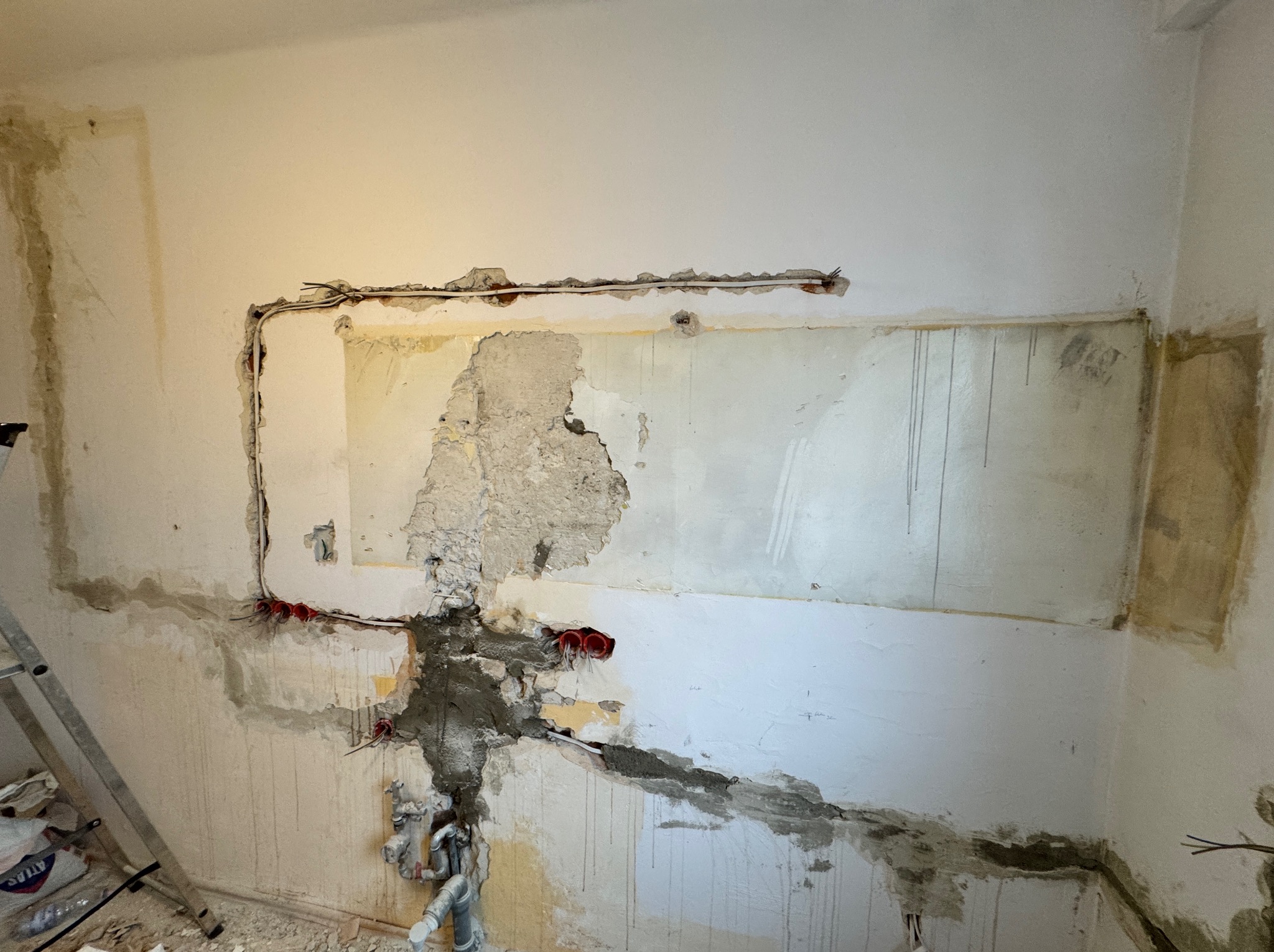 Ściana w trakcie remontu z widocznymi przewodami elektrycznymi w czerwonych puszkach, fragmentami usuniętego tynku i śladami po instalacjach hydraulicznych, drabina malarska w tle.