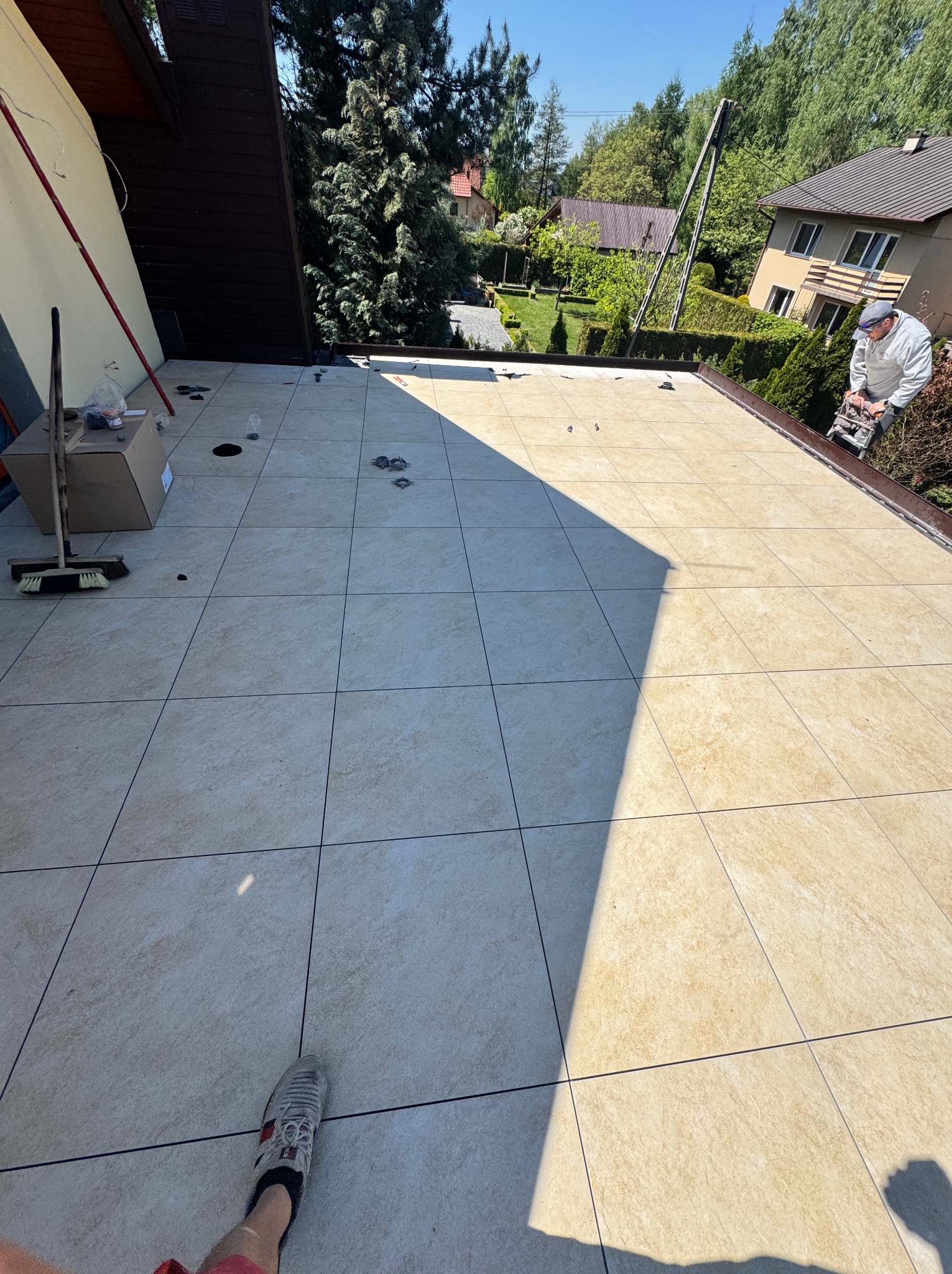 Jasne płytki na tarasie, widoczne narzędzia i pracownik docinający element. Tło: domy i zieleń. Dzień słoneczny, cień na płytkach. Widok z góry, stopa w kadrze.