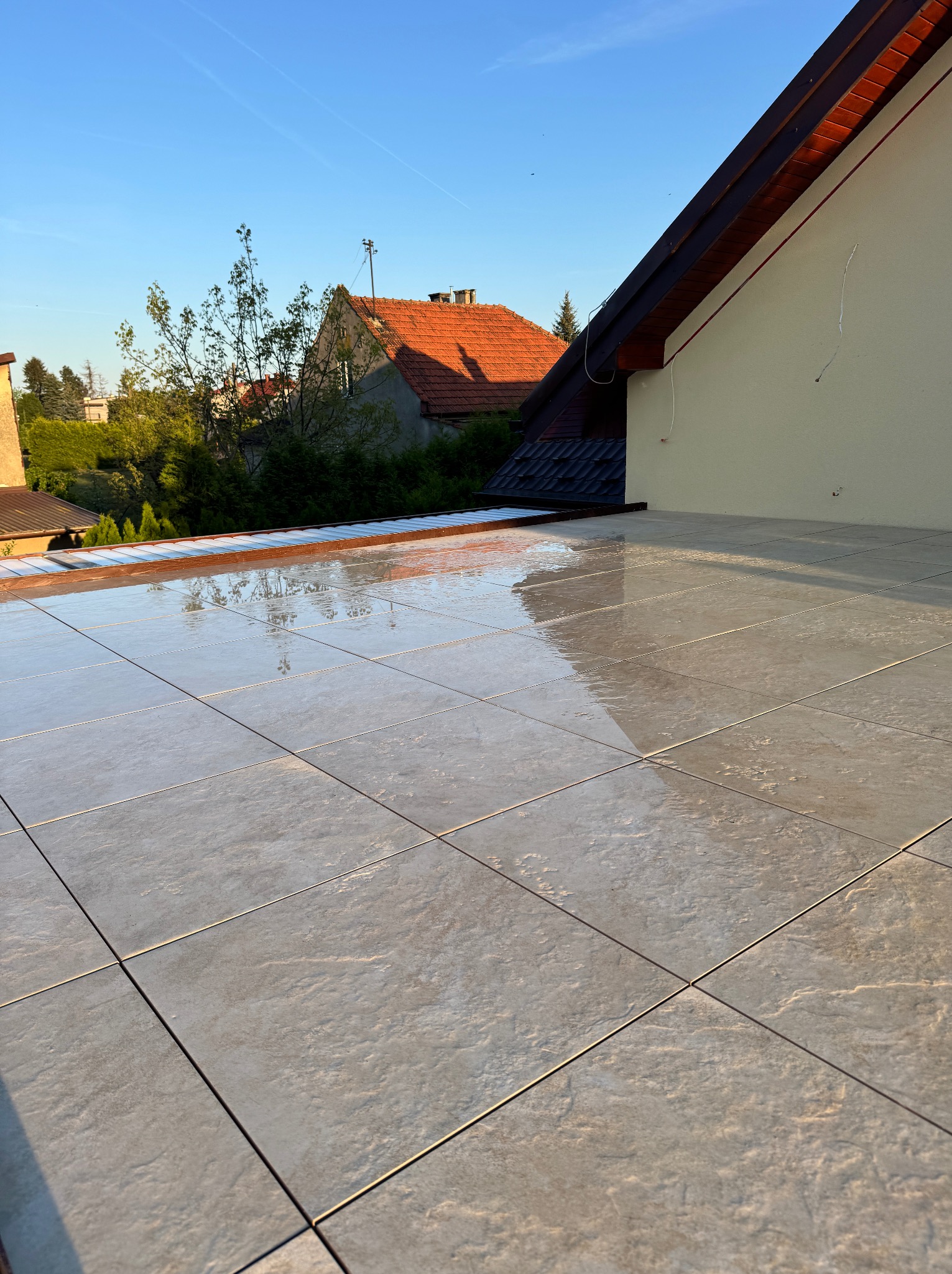 Wyłożony jasnoszarymi płytkami taras z widokiem na domy i zieleń w słoneczny dzień. Płytki odbijają światło, tworząc efekt połysku. Widoczny fragment elewacji budynku.