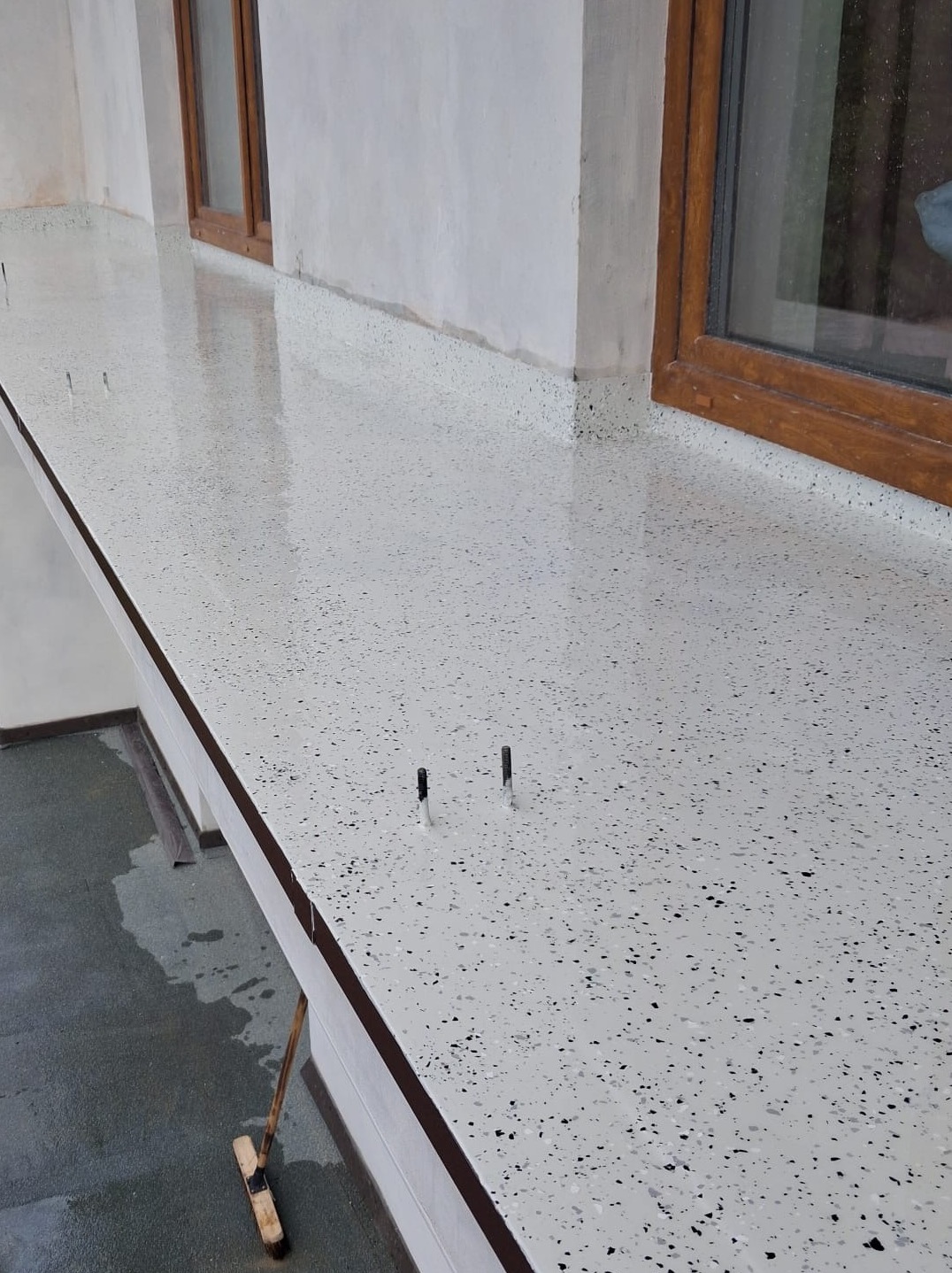 Biała posadzka żywiczna z czarnymi drobinkami, świeżo wylana na balkonie w Krakowie, z widocznymi elementami montażowymi i oknami w tle.