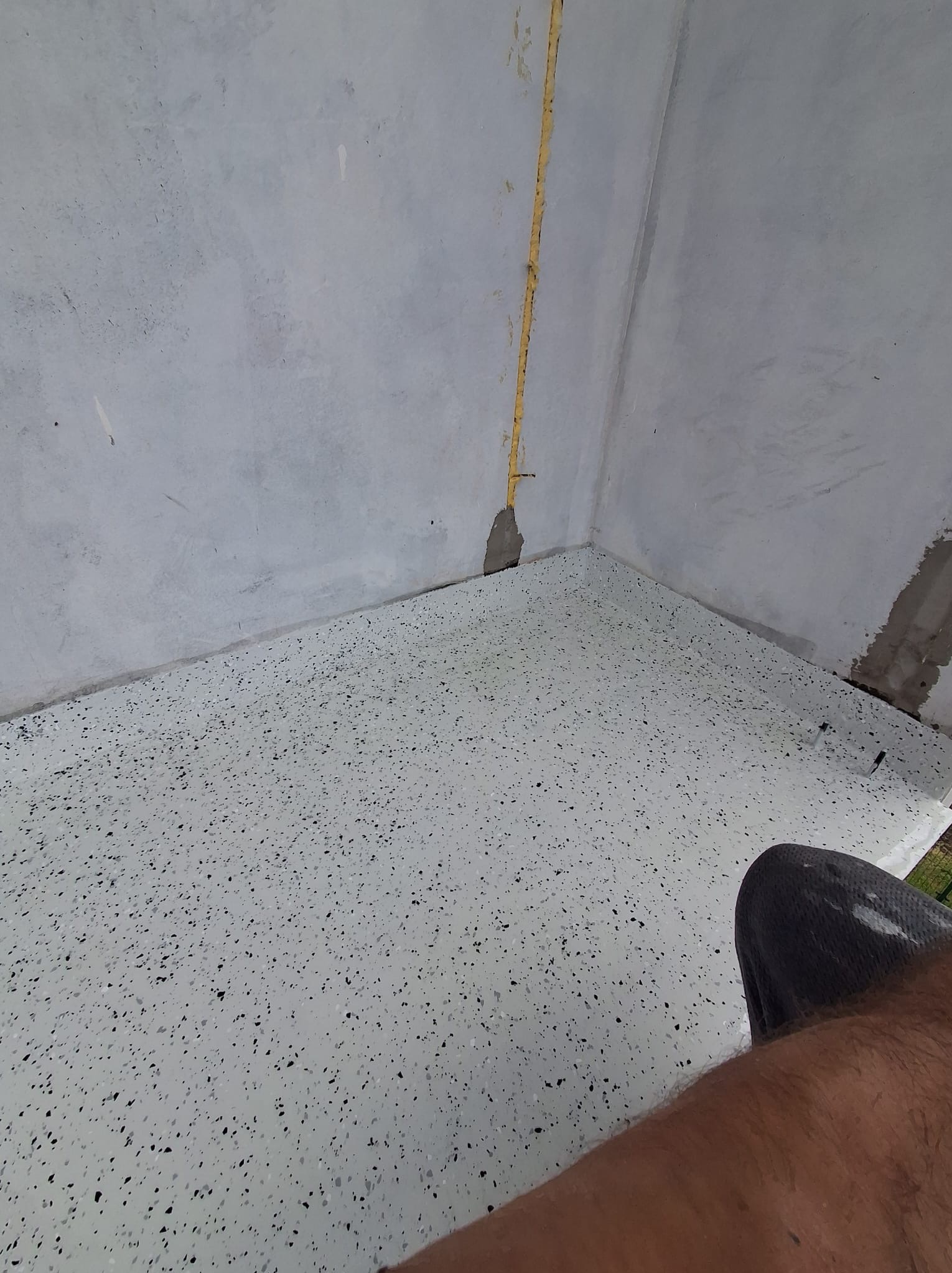 Biała posadzka żywiczna z czarnymi drobinkami w rogu pomieszczenia. Widoczny fragment ściany z żółtą instalacją i fragment nogi osoby.