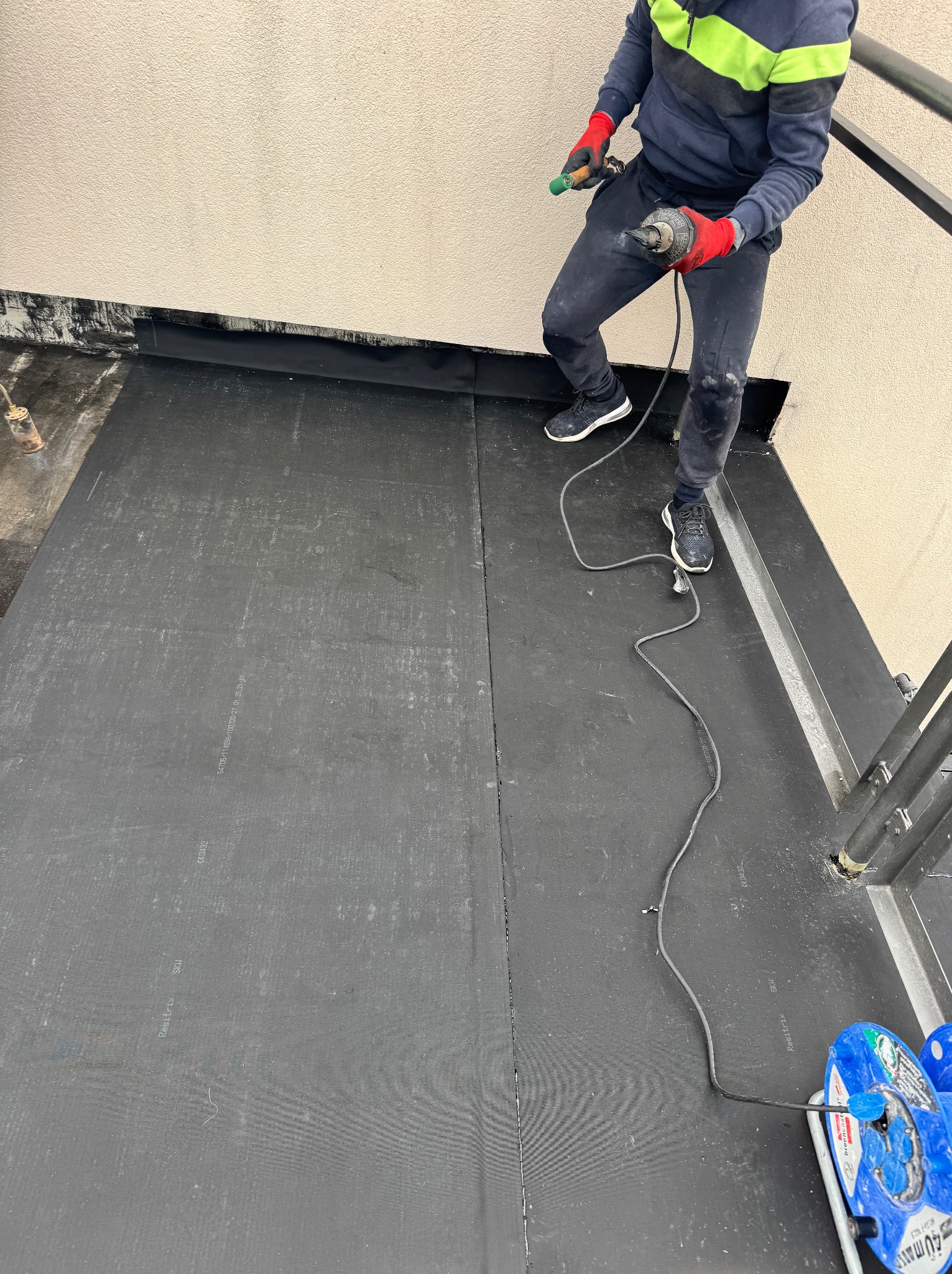 Praca na dachu: dekarz zgrzewa papę termozgrzewalną. Widoczny sprzęt dekarski i fragment barierki. Specyficzny kąt ujęcia podkreśla rozległość powierzchni pokrytej papą.