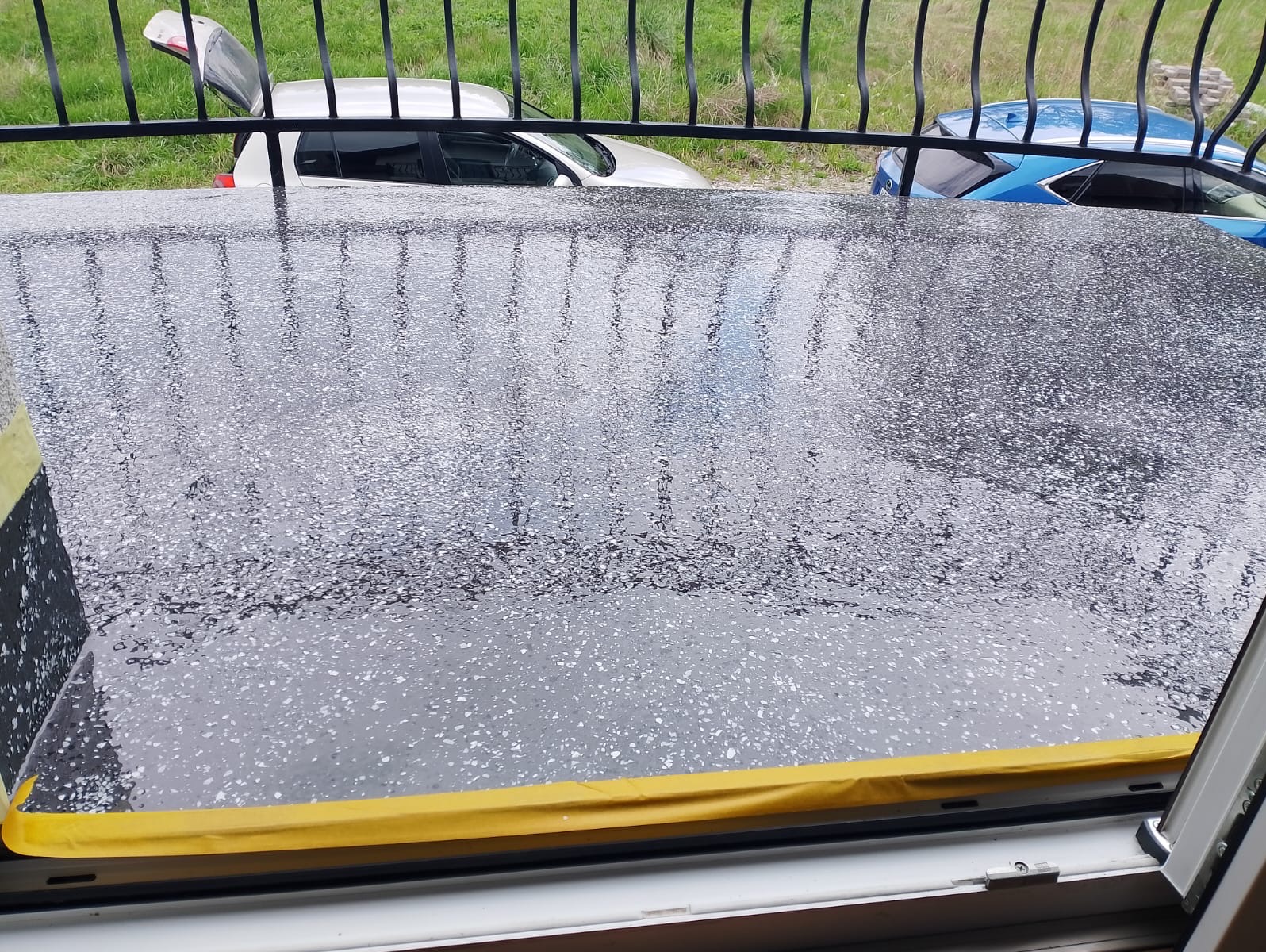 Błyszcząca, ciemnoszara posadzka żywiczna na balkonie po deszczu, zabezpieczona żółtą taśmą malarską przy oknie. W tle barierka i zaparkowane samochody.