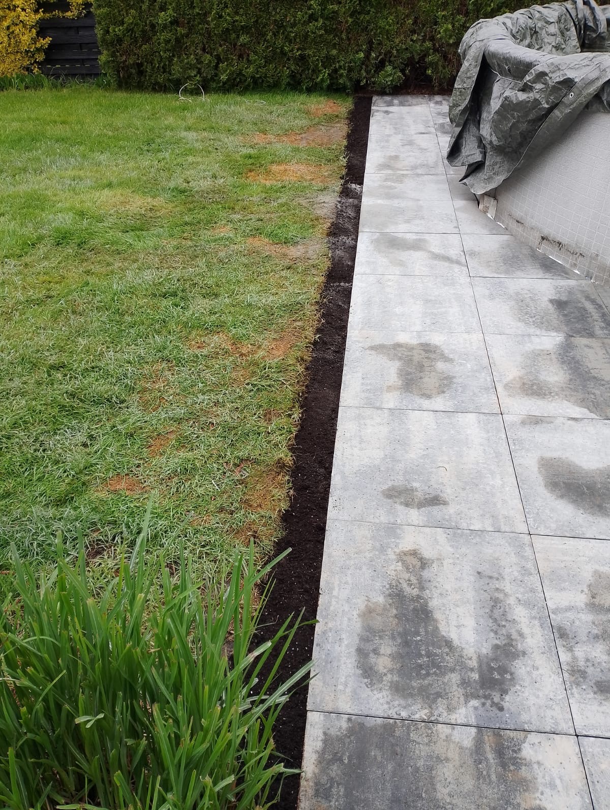 Ułożone płyty tarasowe z szarego betonu, widoczne świeże obrzeże z ziemi, trawa i zieleń w tle. Część tarasu przykryta plandeką. Widok z poziomu gruntu.