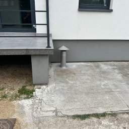 Supply air Bartłomiej Jabłoński - Zewnętrzna instalacja wentylacyjna z metalowym kominem wentylacyjnym przy ścianie domu, widoczny fragment tarasu z szarą balustradą i okno z odbiciem.