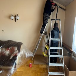 Supply air Bartłomiej Jabłoński - Montaż klimatyzacji: Dwóch monterów na drabinach wierci otwór w ścianie, zabezpieczone meble folią, widoczna lampa robocza i kartonowe pudła.