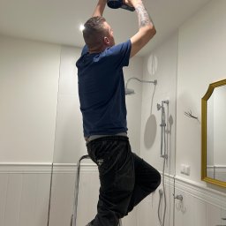 Supply air Bartłomiej Jabłoński - Instalator na drabinie montuje anemostat sufitowy w łazience z białą armaturą i włączonym oświetleniem. Widoczne przewody wentylacyjne.
