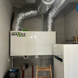 Supply air Bartłomiej Jabłoński - Zmontowana instalacja rekuperacji WANAS z aluminiowymi przewodami wentylacyjnymi w szarej kotłowni. Widoczny niebieski zbiornik wyrównawczy i drabina.