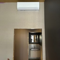 Supply air Bartłomiej Jabłoński - Biała klimatyzacja na ścianie nad wejściem do łazienki z widoczną umywalką i lustrem. Beżowa tapeta i ciemnoszara ściana. Złote wykończenia.