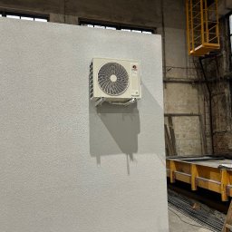 Supply air Bartłomiej Jabłoński - Biała jednostka klimatyzacji marki Gree zamontowana na szarej ścianie o teksturowanej powierzchni w surowym wnętrzu hali z oknami i żółtym podnośnikiem.
