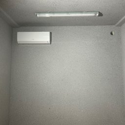 Supply air Bartłomiej Jabłoński - Zamontowany, biały klimatyzator na szarej, teksturowanej ścianie. Nad nim podłużna lampa. W rogu widoczne narzędzia.